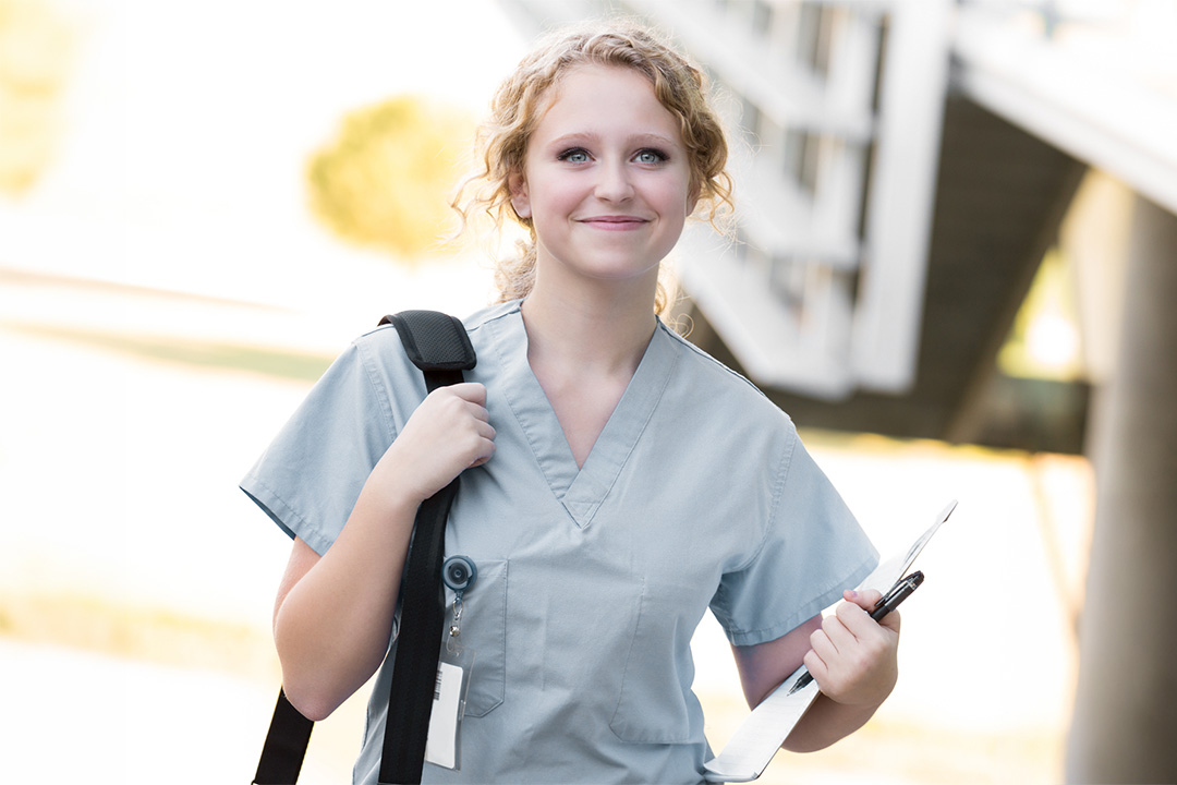 A nursing student wearing scrubs walking forward, holding a clipboard and wearing a backpack on her shoulder.