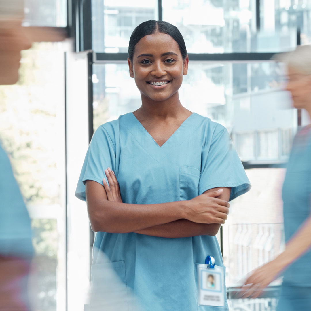 A nurse with her arms crossed, smiling at the camera. There are two nurses walking behind her.