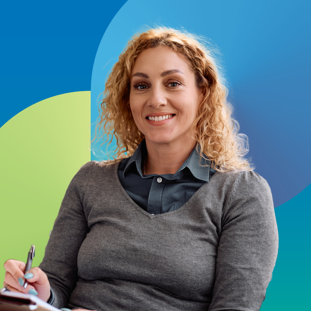 A nurse practitioner smiling at the camera while seated and writing on a clipboard with a pen.
