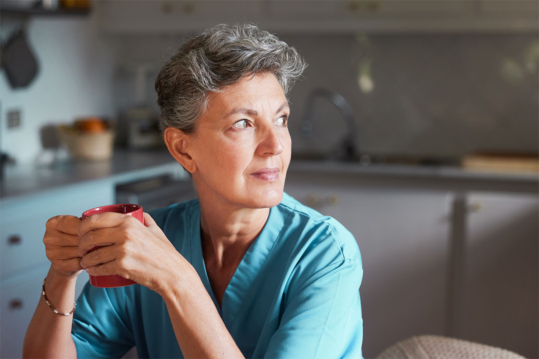 Une infirmière blanche âgée portant une blouse, tenant une tasse de café et regardant attentivement par la fenêtre de sa maison.