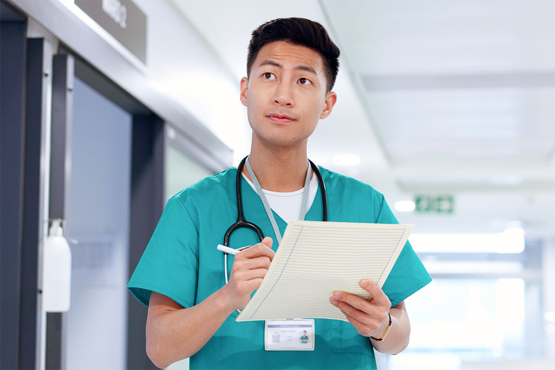 Un infirmier d’Asie de l’Est portant une blouse dans un couloir d’hôpital, tenant des documents et perdu dans ses pensées.